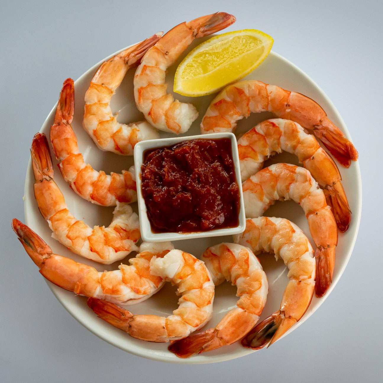 Shrimp cocktail arranged in a ring around the edge of a crystal serving platter with cocktail sauce and a lemon wedge.