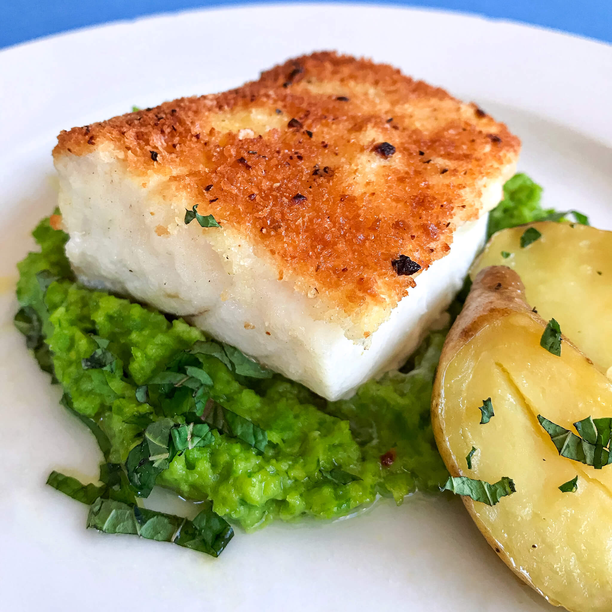 panko crusted pan fried 5 ounce cod portion on a bed of mushy peas with potato wedges on the side served on a white plate