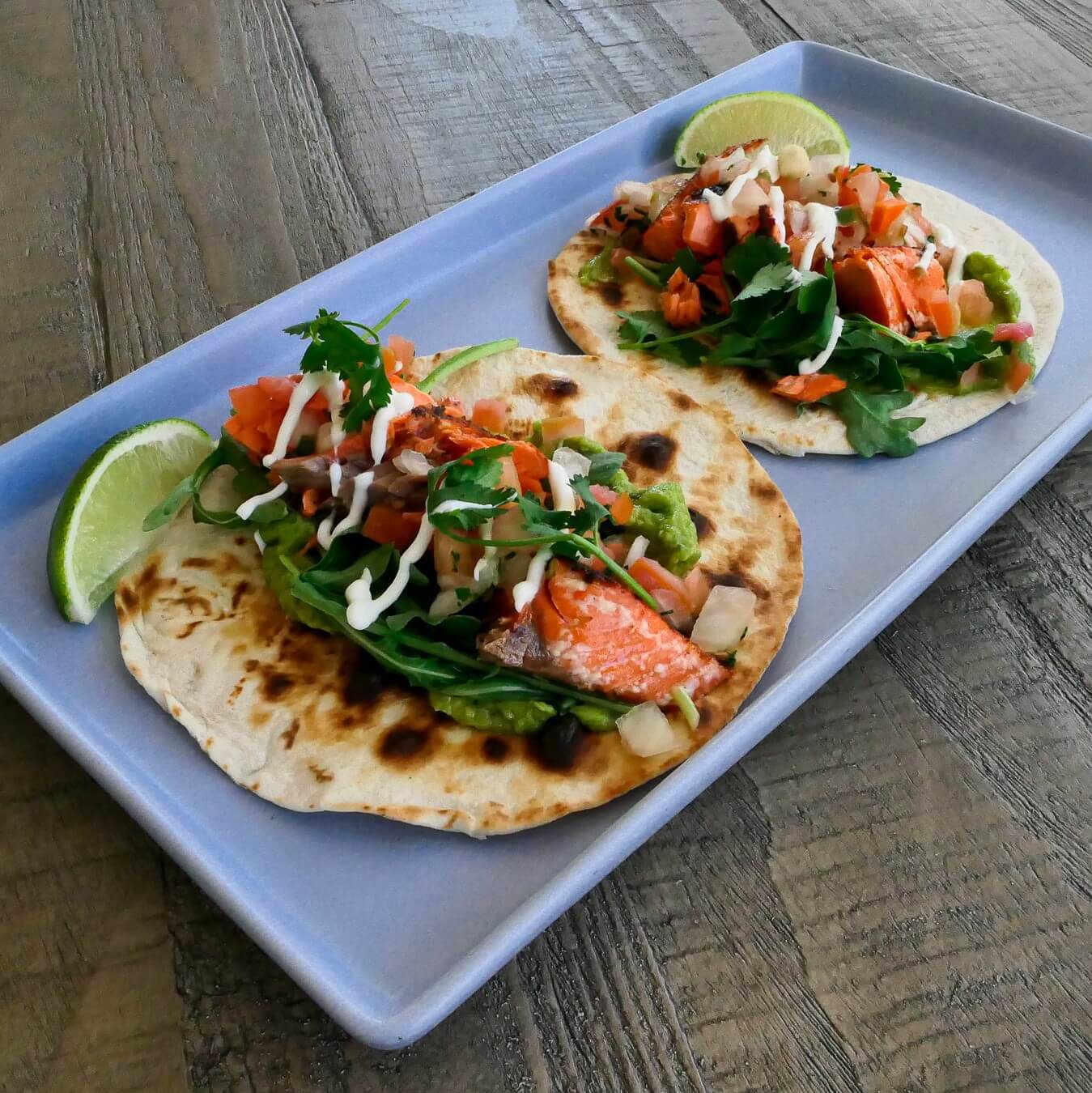 Two wild salmon tacos with baby arugula, salsa and sour cream on a plate with lime wedges on the side