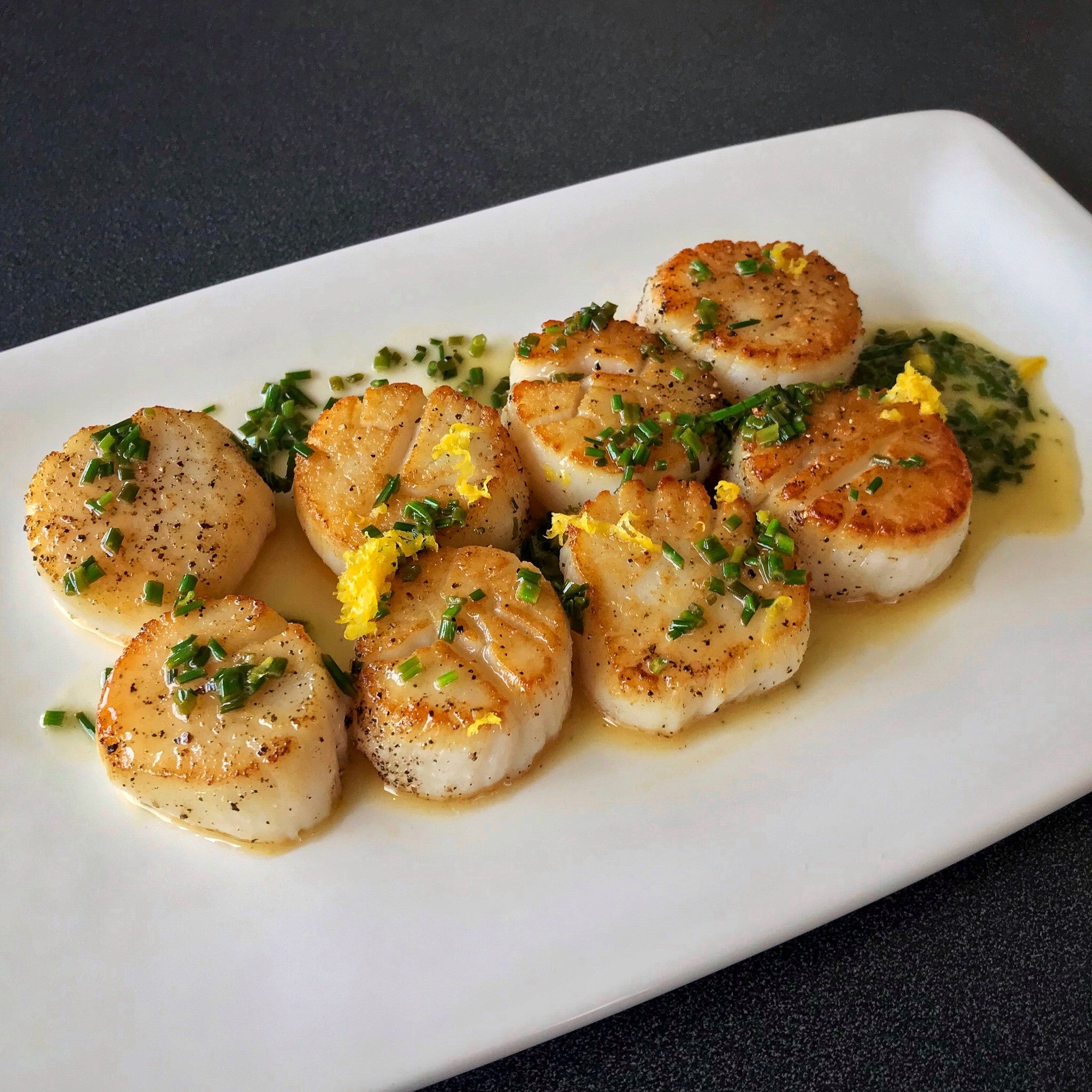 pan seared scallops topped with lemon butter sauce, chopped chives, and lemon zest, served on a white plate