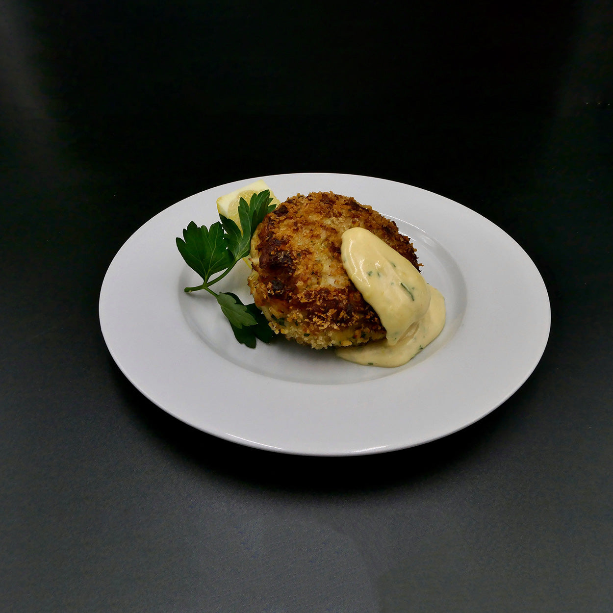 pan fried homemade crab cake topped with a dollop of spicy aioli sauce and a sprig of fresh parsley served on white plate with black background
