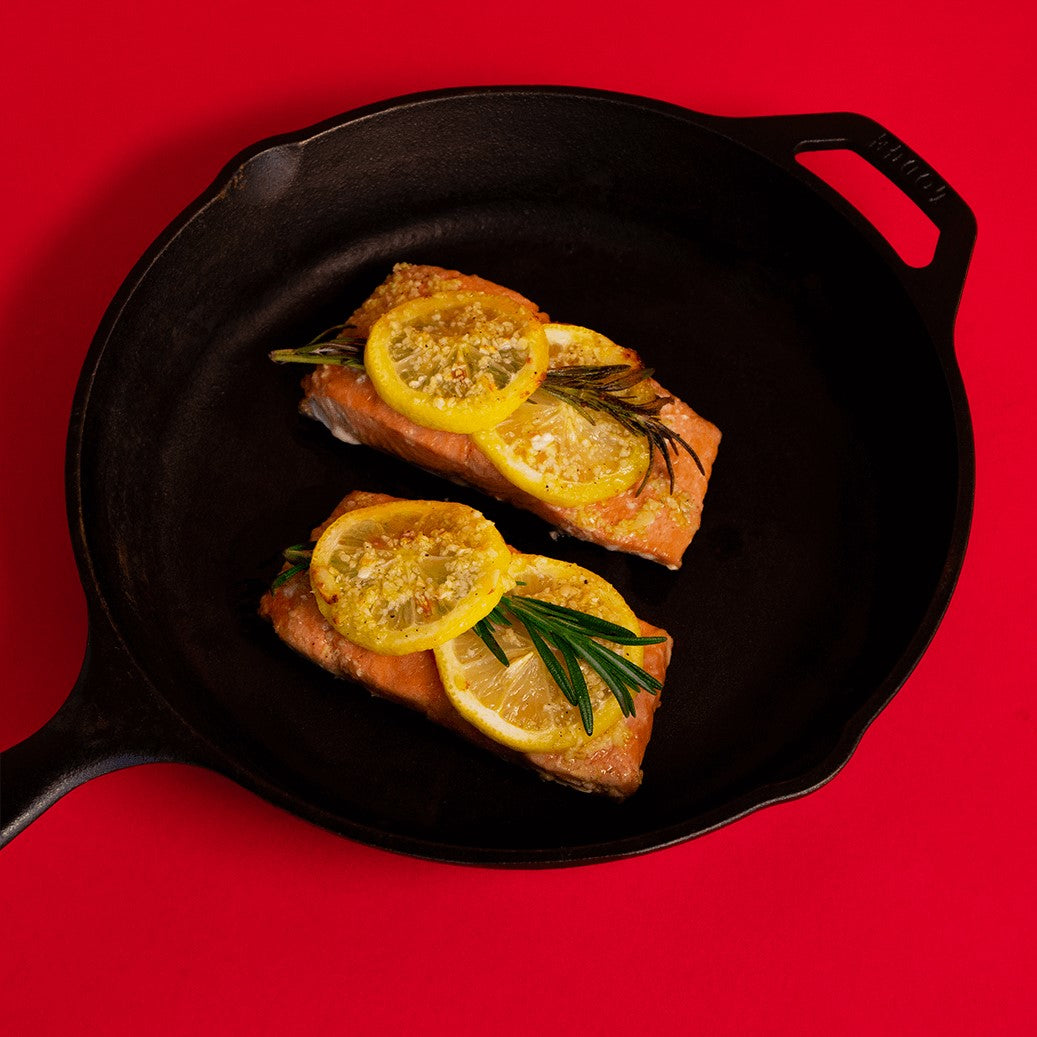 two wild salmon portions in a cast iron pan topped with rosemary sprigs and lemon slices on a red background