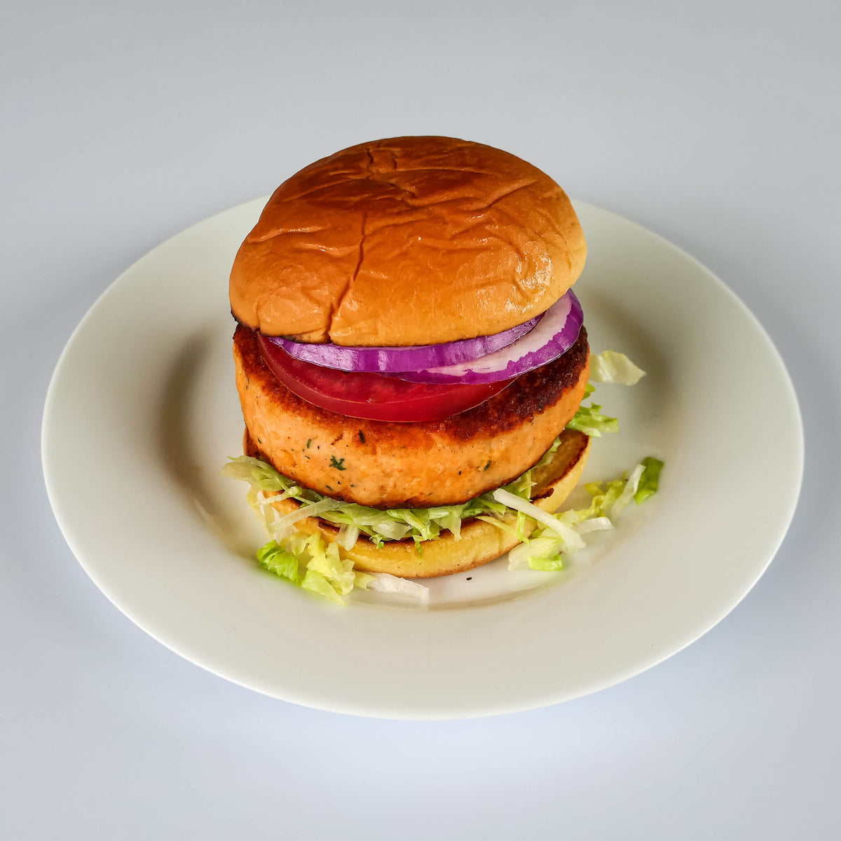 pan fried salmon burger on a bun with lettuce, tomato, and onion served on a white plate